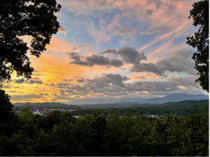 Sunset view over a forested valley with scattered clouds and mountains in the distance, framed by trees on both sides